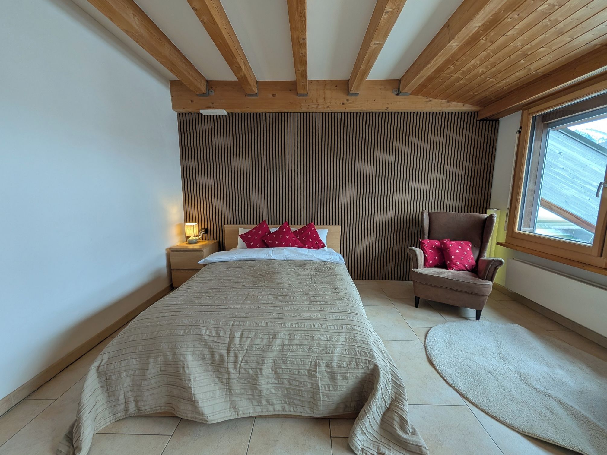 Un dormitorio con cama, mesita de noche, una silla, ventana y techo de madera.