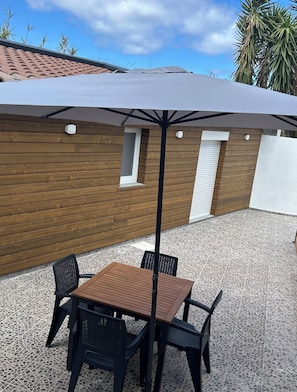 Een terras buiten met een tafel, stoelen, een grijze parasol en een houten muur met ramen.