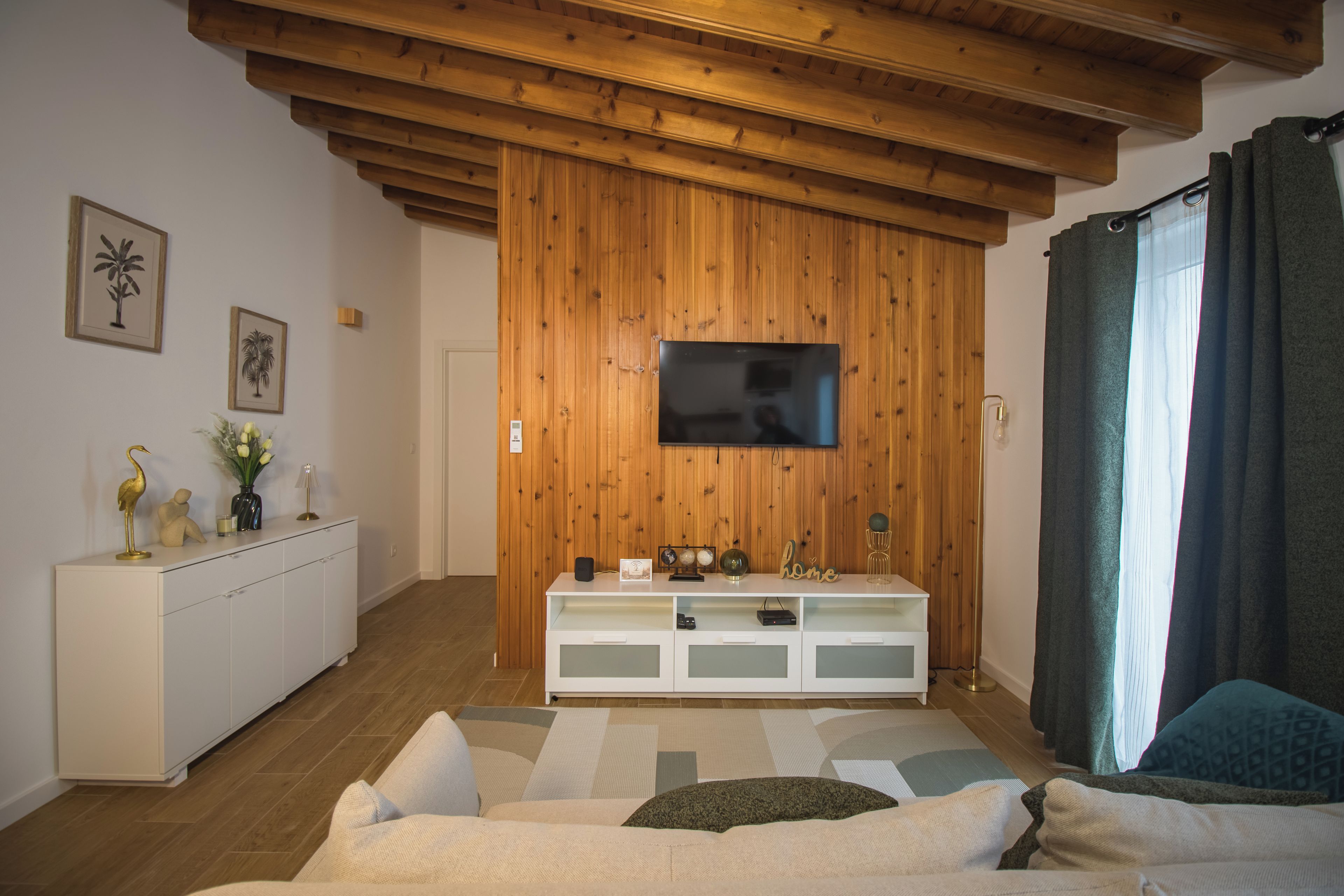 Un salon moderne avec un mur recouvert de panneaux de bois, un téléviseur à écran plat fixé au-dessus d’un meuble de télévision blanc, un canapé et une grande fenêtre agrémentée de rideaux.