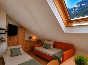 A compact room with a sloped ceiling, a sofa bed, a television mounted on a wooden cabinet, and a window with a mountain view. - Oria Dolomites Stay (Alba)
