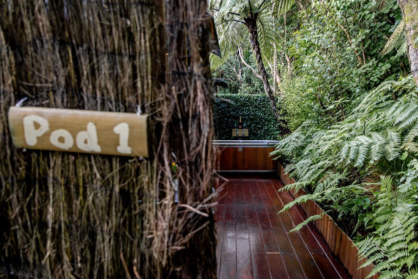 A wooden pathway leading to a hot tub labeled 'Pod 1' surrounded by lush greenery.
