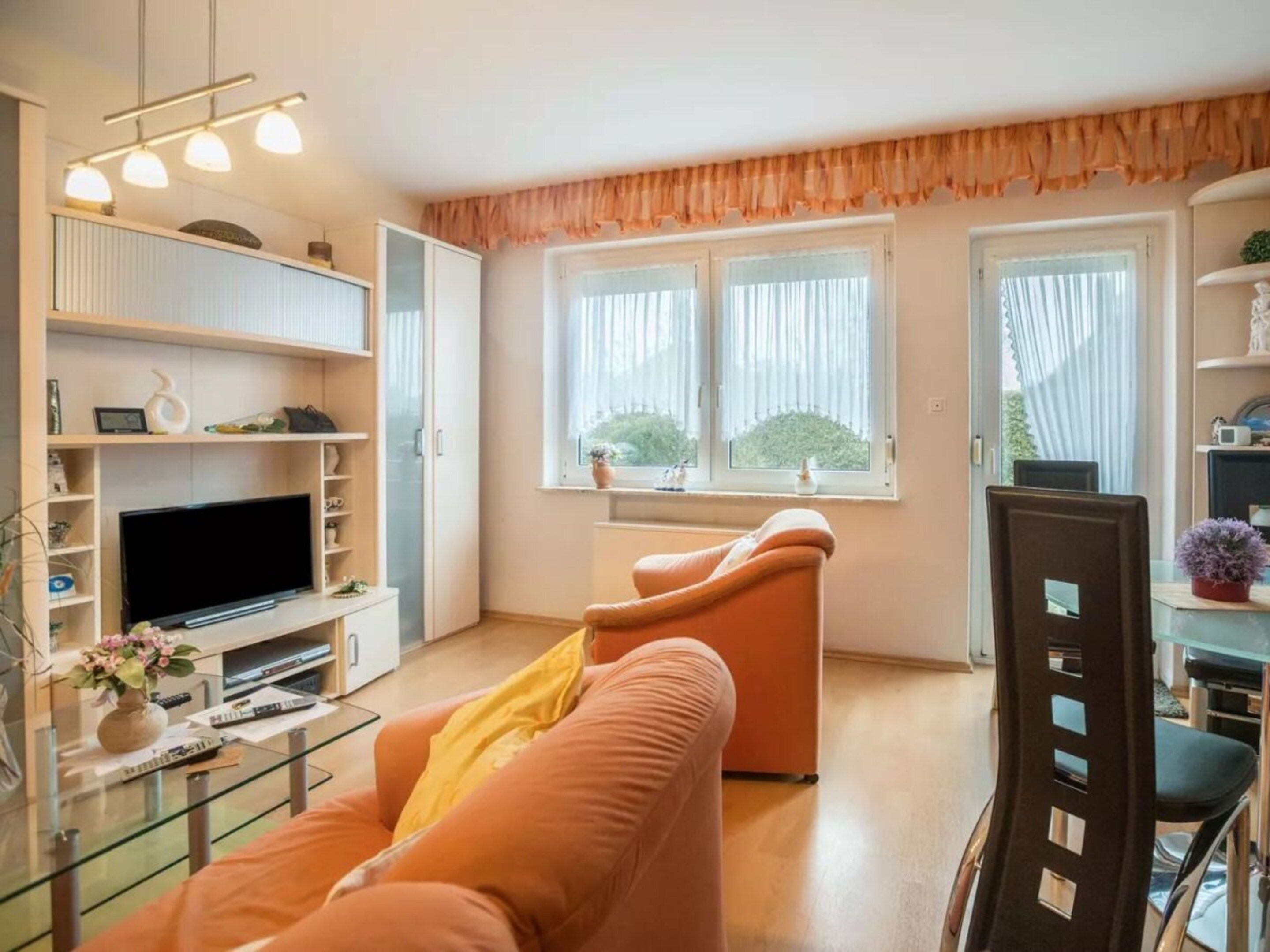 A modern living room with a sofa, TV, and glass coffee table. There are shelves with decorative items and a window with curtains.
