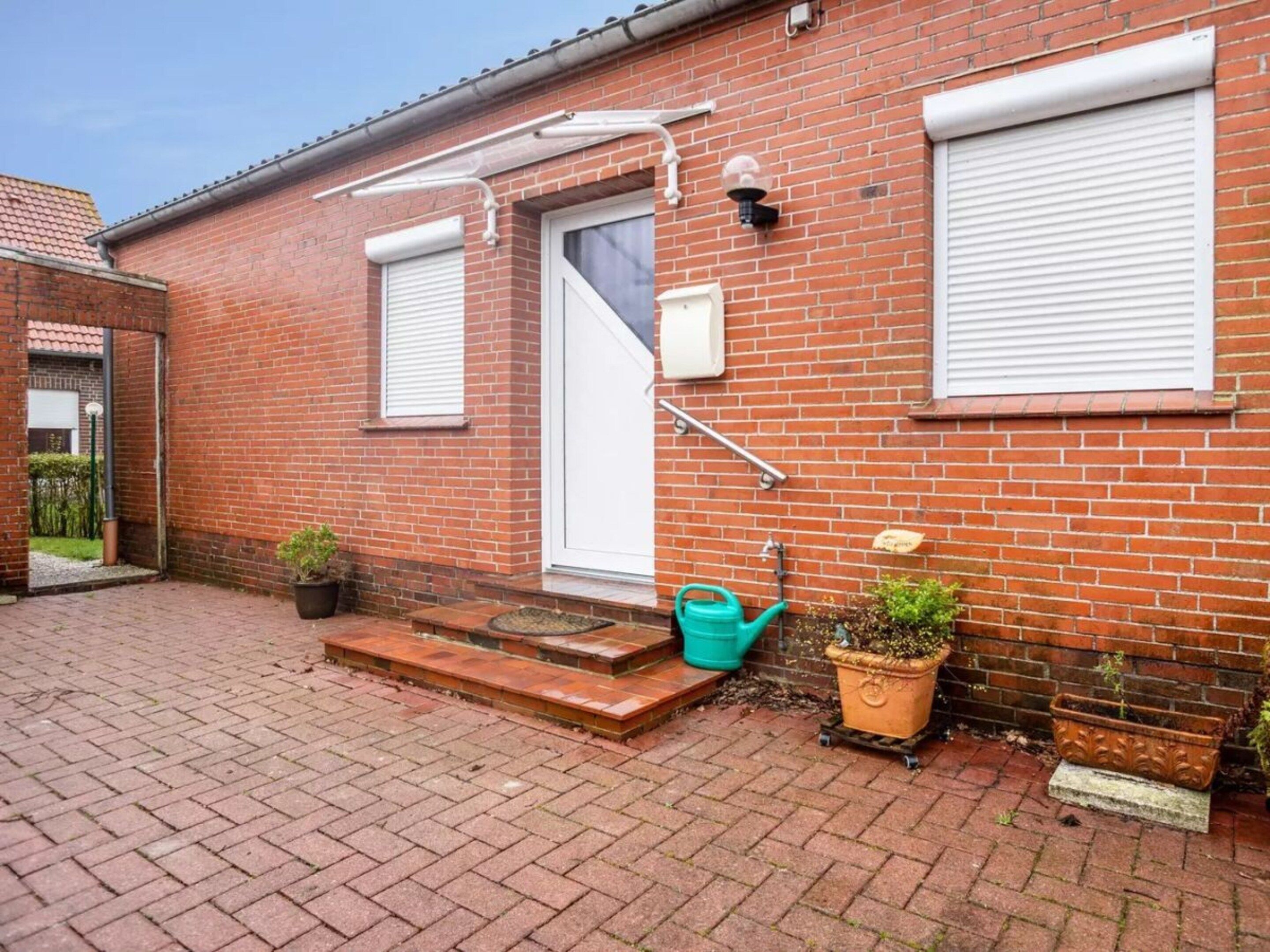 Een bakstenen huis met een witte deur, een klein portaal met treden en een groene gieter.