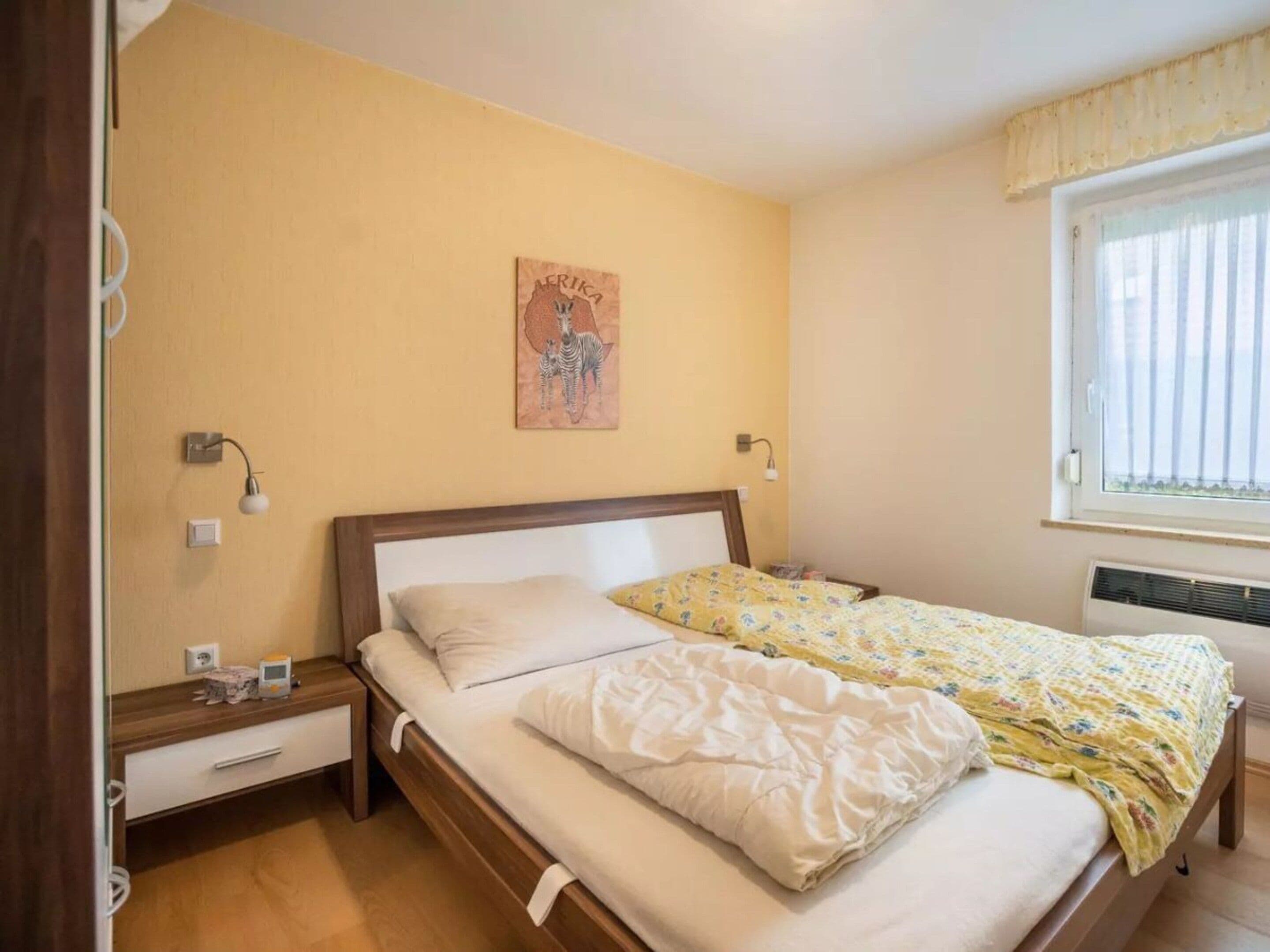 A bedroom with a bed, bedside tables, a wall-mounted lamp, a window with curtains, and a wall painting.