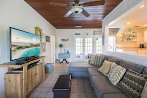 A living room with a television, a sofa, a ceiling fan, and a kitchen area visible through an open door. - Private 3BR Pool Home | Pet Friendly | Sleeps 8 (Bradenton)