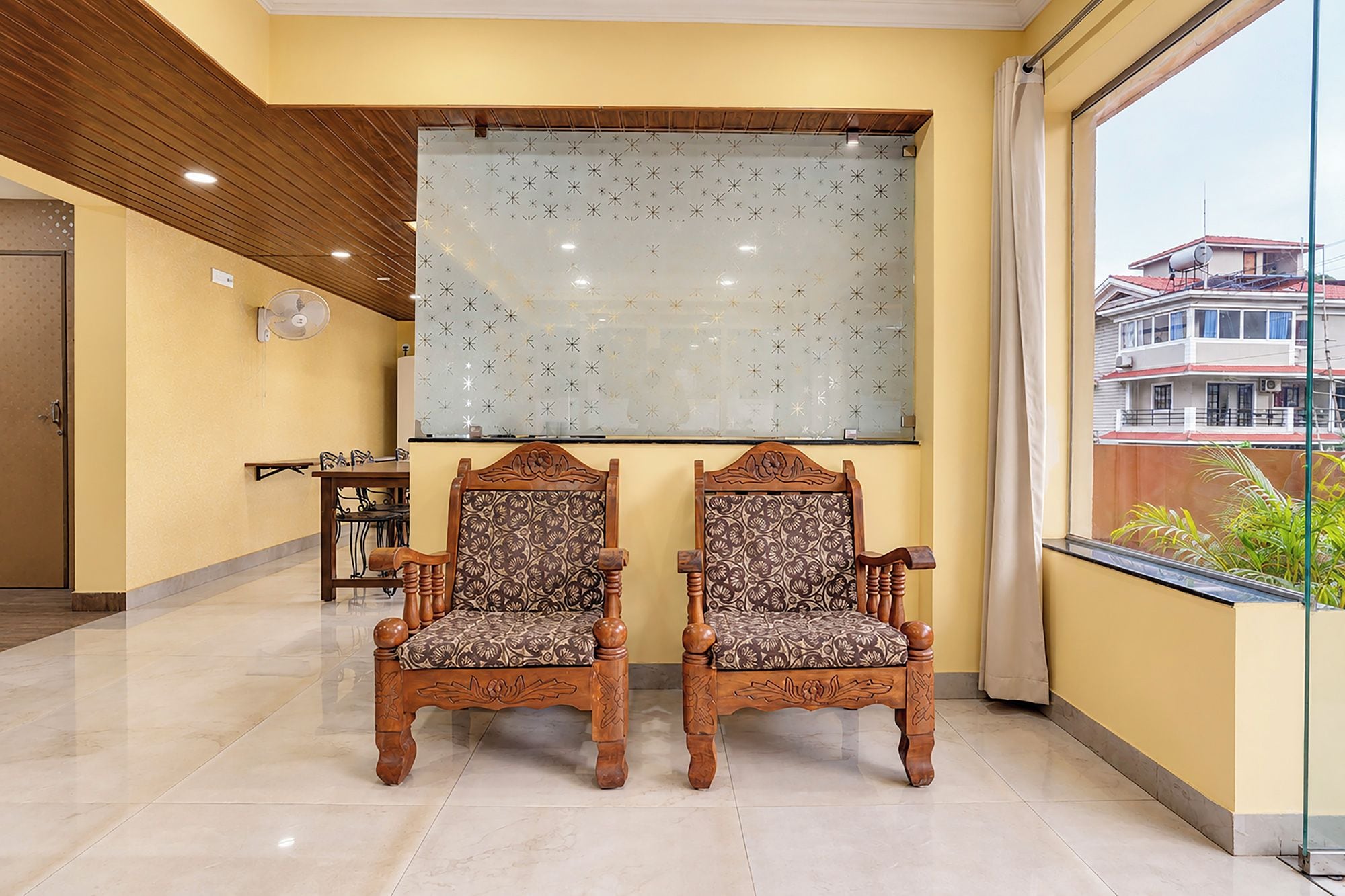 Quarto com duas cadeiras elaboradas, uma janela ampla com vista para edifícios e um painel decorativo fixado na parede.