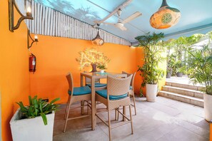 Un espacio exterior para comer con sillas de mimbre y una mesa, rodeado de plantas en macetas y una pared decorativa.