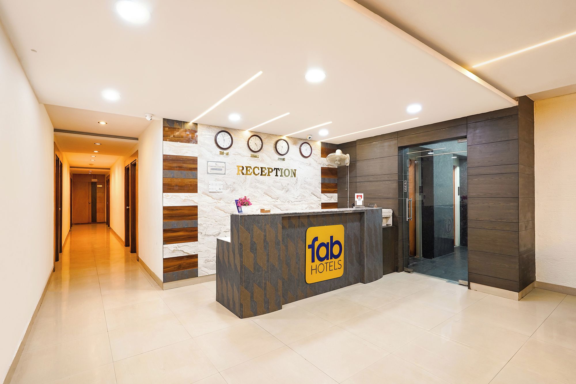 Zona de recepción de hotel con mostrador, relojes y puerta de vidrio que da al interior.