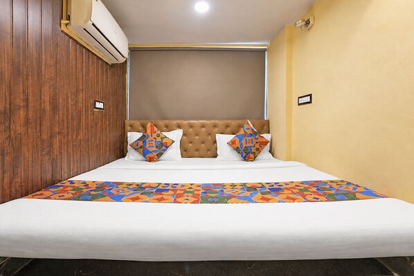 A hotel room with a bed, a headboard, a wall-mounted air conditioner, and a wall socket.