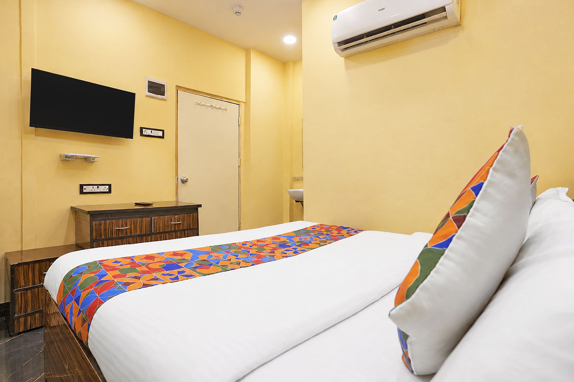 Una habitación de hotel con una cama, un televisor montado en la pared, una mesita de noche y un aire acondicionado.