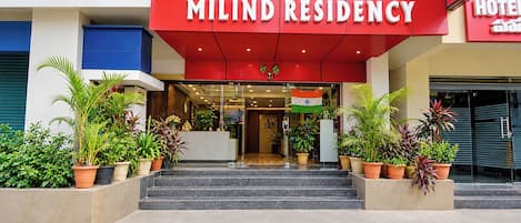 La entrada del Hotel Milind Residency, con fachada roja, puertas de cristal y plantas en macetas.