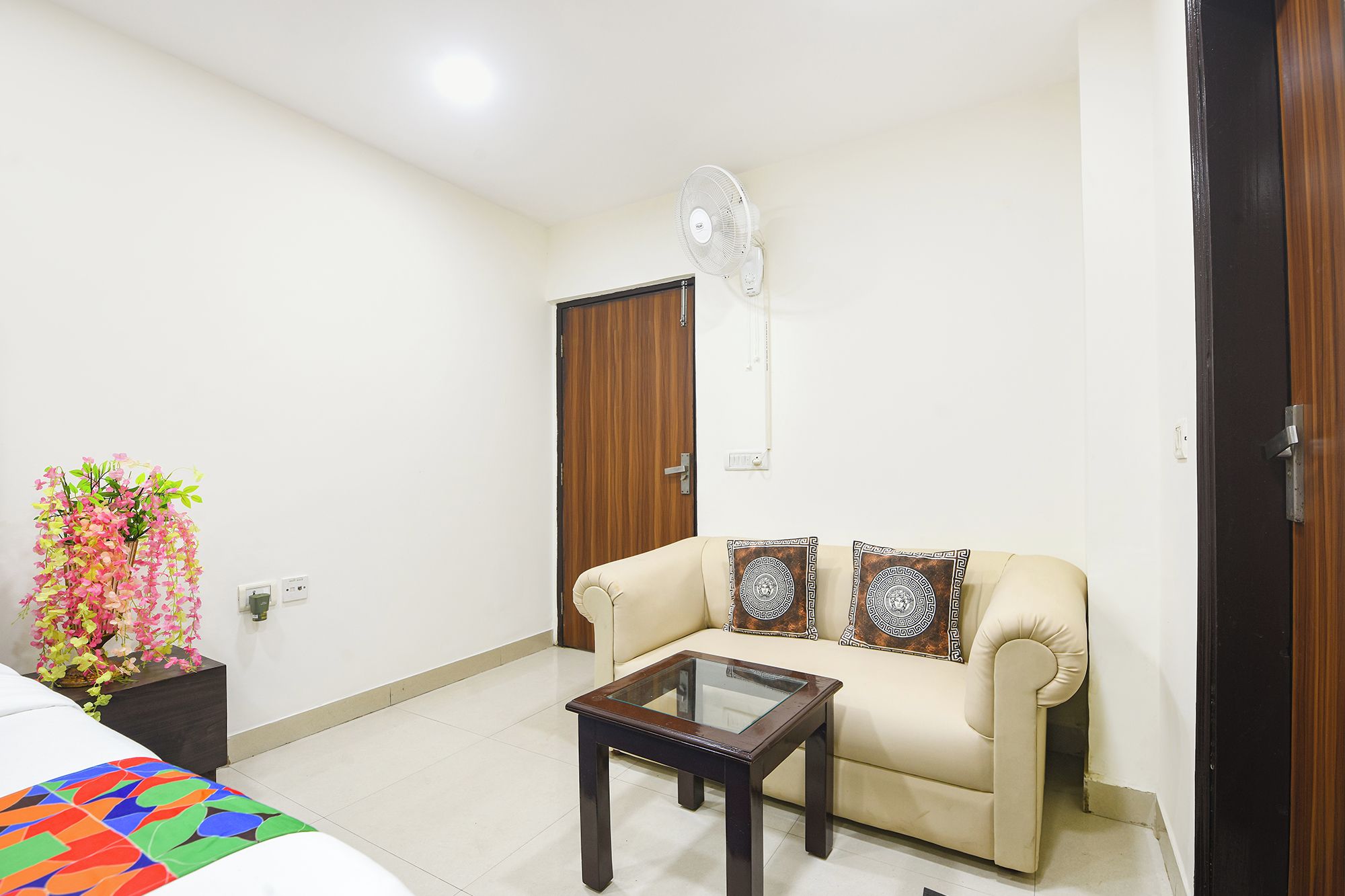 A living room with a sofa, a glass-top coffee table, a wall-mounted fan, and a decorative plant.