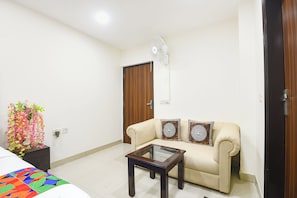 A living room with a sofa, a glass-top coffee table, a wall-mounted fan, and a decorative plant. - FabHotel Sbs Palza (New Delhi)
