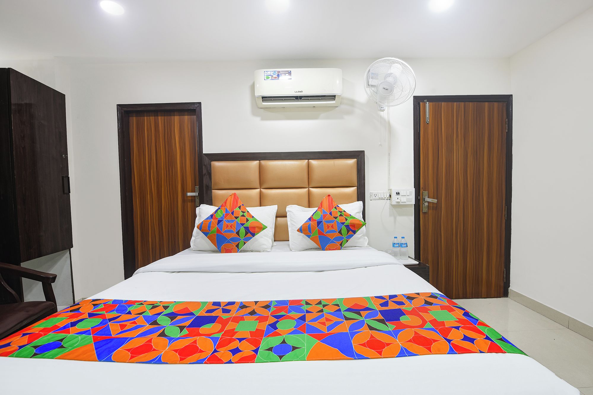 A hotel room with a bed, two pillows, a bedside table, an air conditioning unit, a wall-mounted fan, and wooden doors.