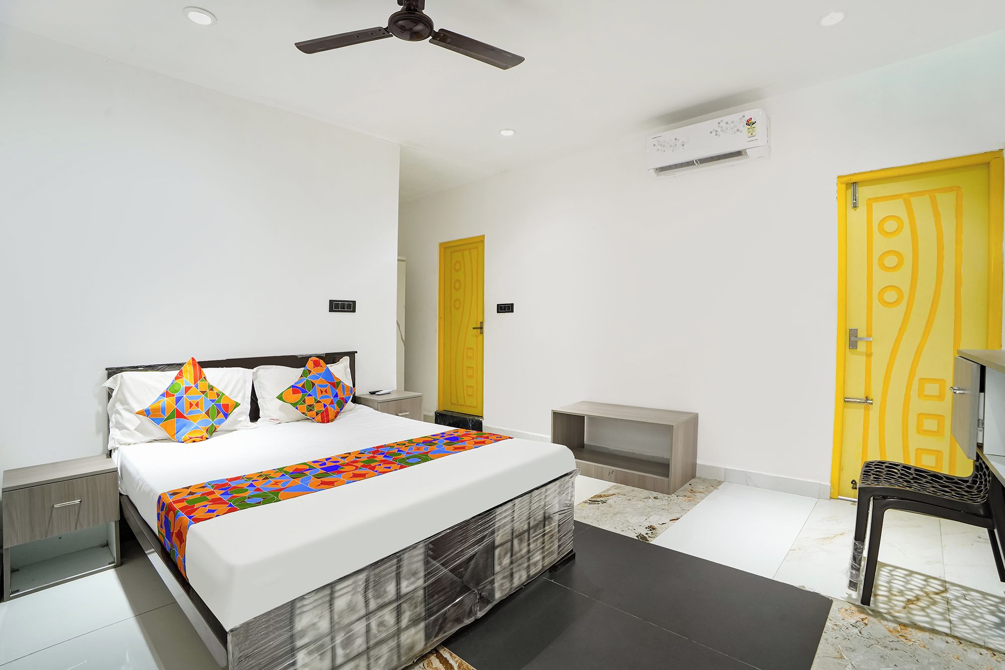 A modern bedroom with a bed, bedside tables, a ceiling fan, an air conditioning unit, and a yellow door.