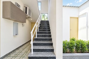 Una escalera con barandilla que lleva a un piso superior, un equipo de aire acondicionado instalado en la pared, una puerta y un pequeño jardín con plantas.