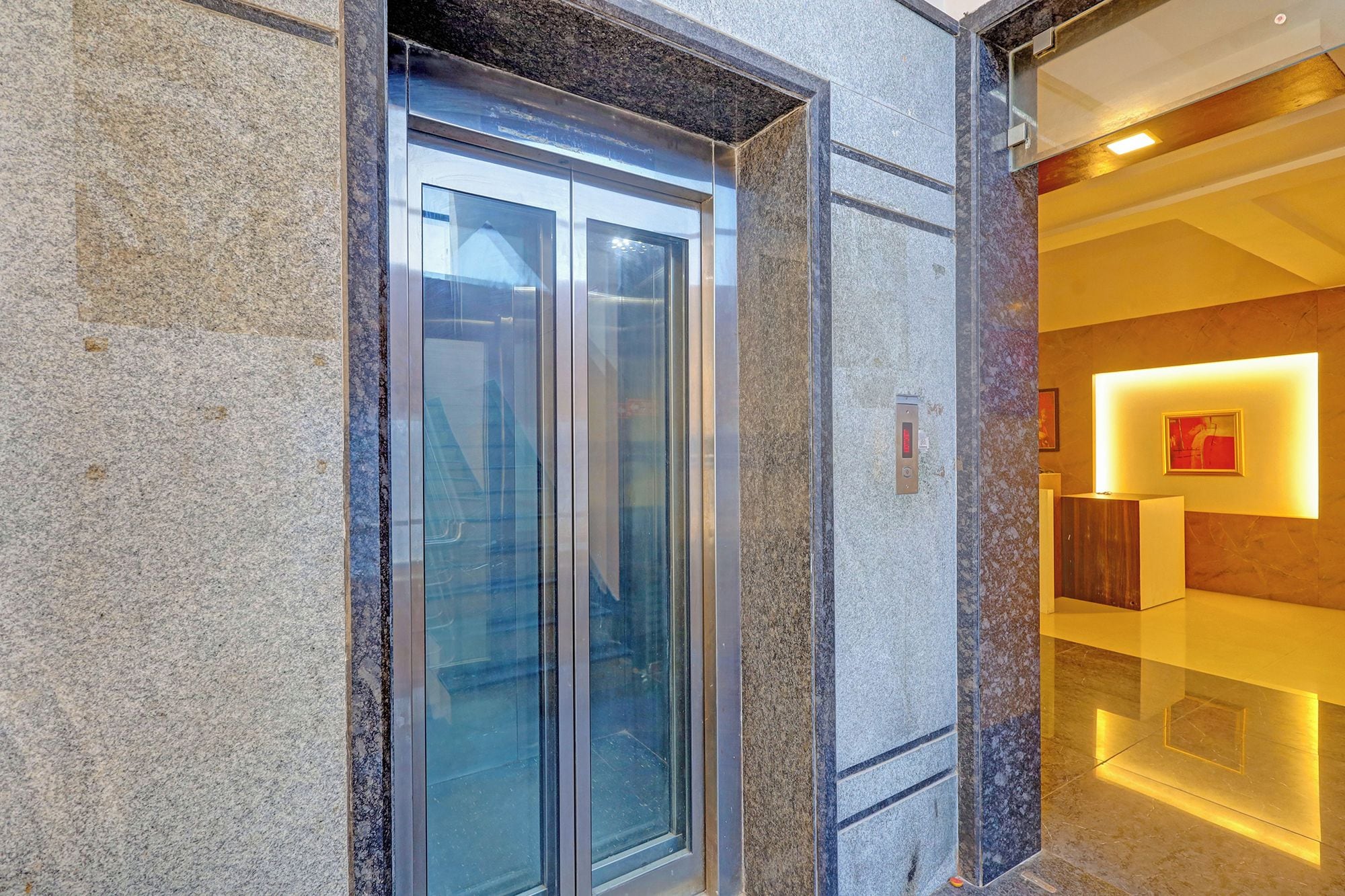 Un ascensor con puertas de vidrio, pared de mármol e interior moderno con una pared de acento rojo y un cuadro enmarcado.