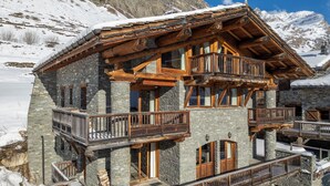 Un chalet de varios pisos con paredes de piedra y balcones de madera, con una imponente montaña nevada de fondo.
