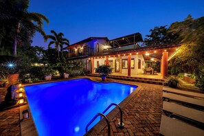 A swimming pool with a clear blue water surface, surrounded by a brick patio, and a building with a balcony in the background. - Villa Brisas in Coral Gables area (Miami)