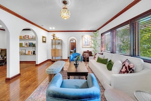 A spacious living room with a blue armchair, white sofa, and a wooden coffee table. There is a bookshelf, a mirror, and a window with blinds. - Villa Brisas in Coral Gables area (Miami)
