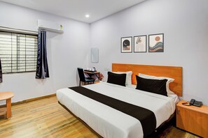Habitación de hotel con cama, escritorio, silla, mesita pequeña, mesita de noche y espejo montado en la pared.