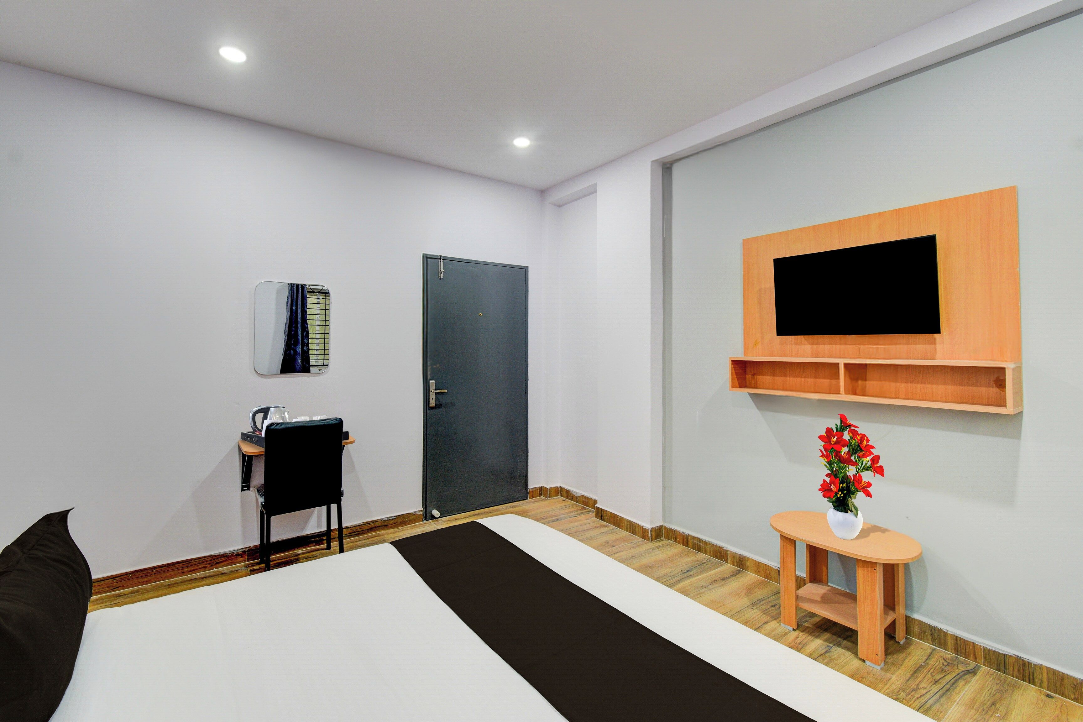 A hotel room with a bed, a desk with a chair, a TV mounted on the wall, and a small table with a vase of flowers.