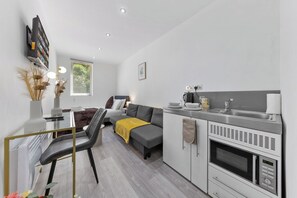 A modern kitchenette with a microwave, stove, and sink. There is a sofa and a chair in the living area, and a bed with a headboard.