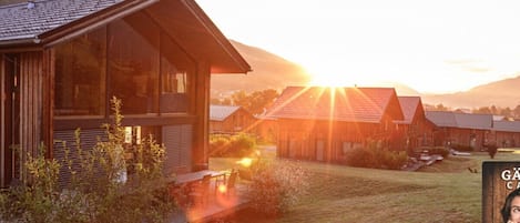 A wooden cabin with a deck, surrounded by a grassy area and mountains in the background.