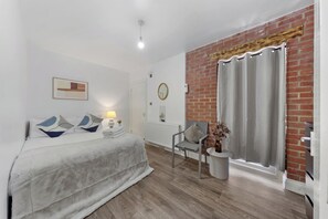 A bedroom with a bed, a chair, a small table, a mirror, and a brick wall.