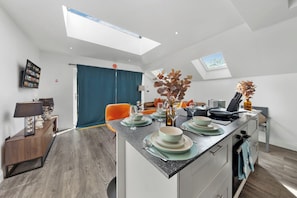 A modern kitchen with a central island, a dining area with orange chairs, and a large window providing natural light.