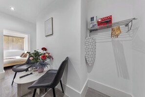 A modern bedroom with a bed, a bedside table with a remote control, a chair, a potted plant, and a wall-mounted shelf with a box and towels.
