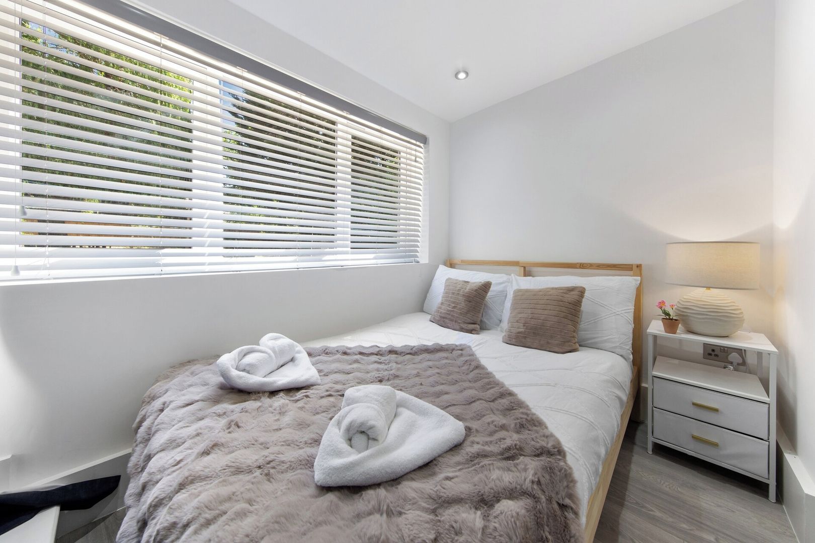 Quarto moderno com cama grande, roupa de cama branca, tapete felpudo, mesa de cabeceira com abajur e janela com persianas.