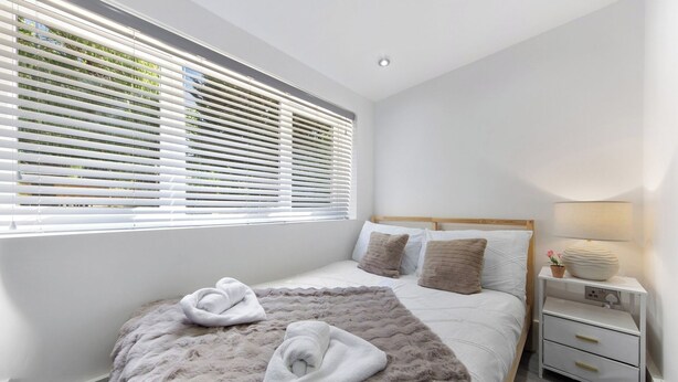 Un dormitorio moderno con una cama grande, ropa de cama blanca, una alfombra mullida, una mesita de noche con lámpara y una ventana con persianas.