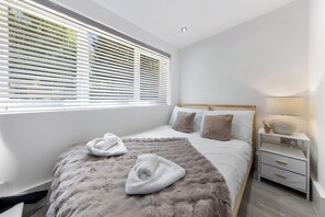 A modern bedroom with a large bed, white bedding, a fluffy rug, a bedside table with a lamp, and a window with blinds.