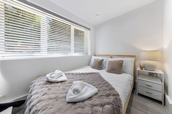 A modern bedroom with a large bed, white bedding, a fluffy rug, a bedside table with a lamp, and a window with blinds.