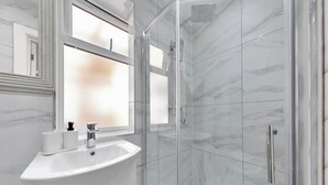 Un baño moderno con ducha envuelta en cristal, un lavabo blanco con un espejo encima y paredes con diseño de mármol.