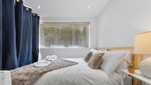 Una cama bien hecha con una manta suave, dos almohadas y una mesita de noche con una lámpara y una planta en maceta.