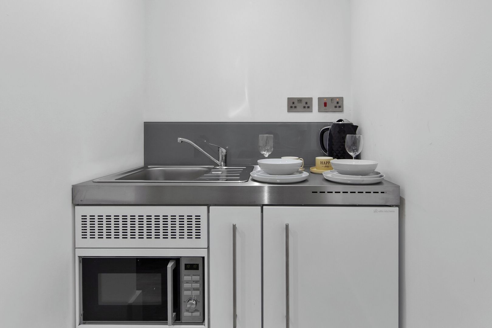 Un angolo cottura compatto con lavello in acciaio inox, forno a microonde e macchina da caffè.