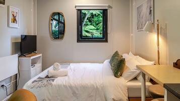 A hotel room with a bed, a desk, a television, a window with a view of greenery, and artwork on the walls.