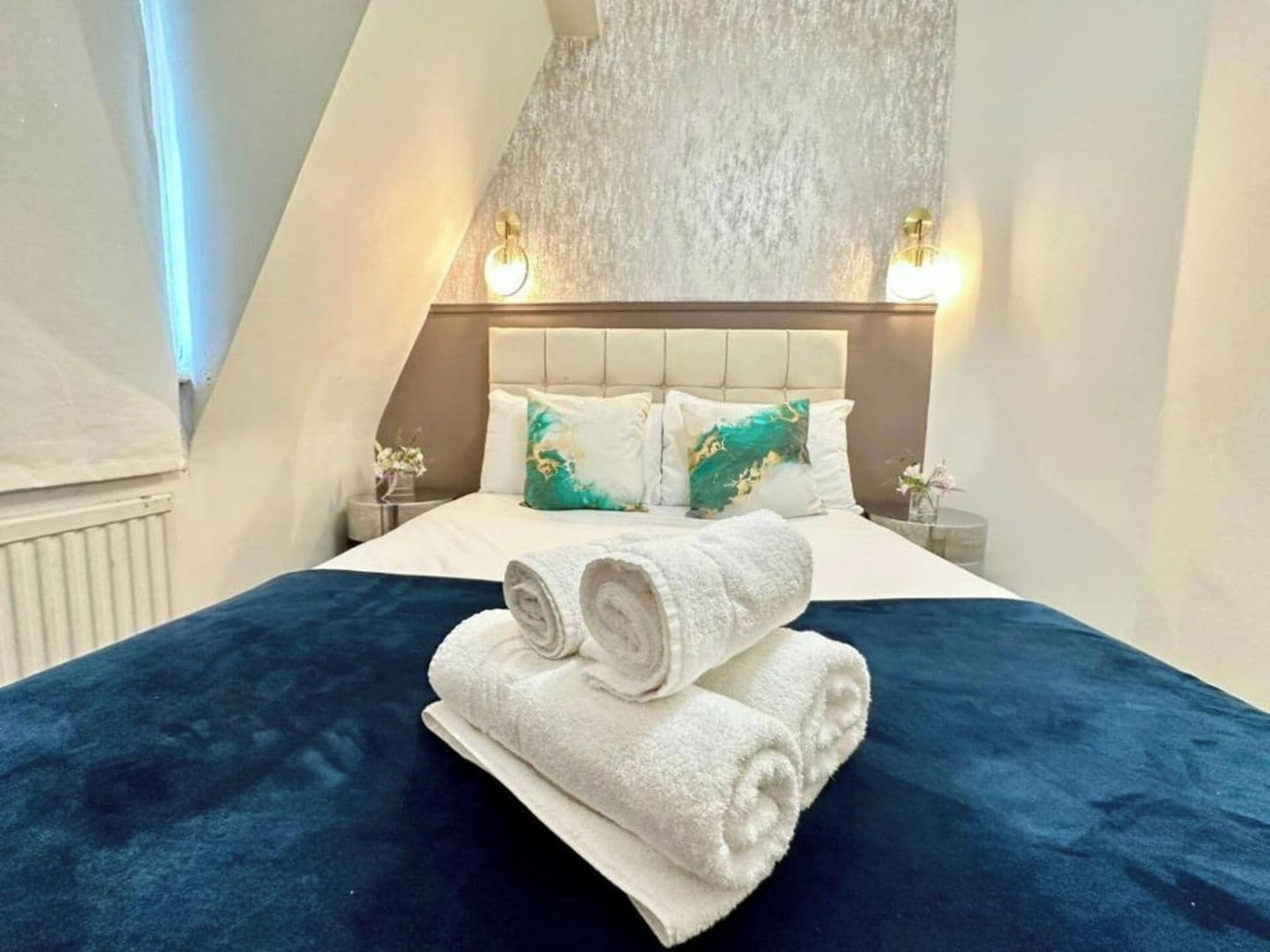 Quarto de hotel com cama, duas mesas de cabeceira e luminária fixada na parede.