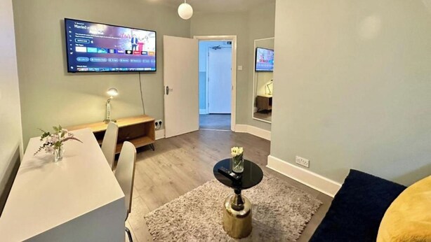Una sala moderna con un televisor de pantalla plana montado en la pared, una zona de comedor con una mesa y sillas blancas, y un pasillo que conduce a otra habitación.