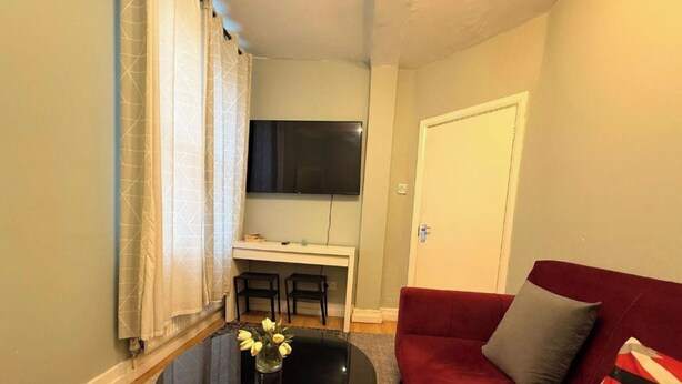 Un espacio habitable compacto con un sofá rojo, una pequeña mesa redonda con un jarrón de flores, una zona de comedor con dos sillas y un televisor montado en la pared.