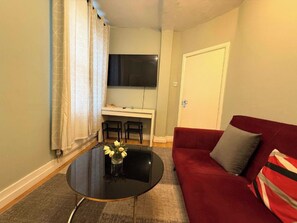 A compact living space with a red sofa, a small round table with a vase of flowers, a dining area with two chairs, and a TV mounted on the wall. - Central London Apartment (Greater London)