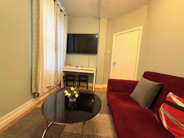 En kompakt opholdsstue med en rød sofa, et lille rundt bord med blomster, et spiseområde med to stole og et fjernsyn ophængt på væggen.