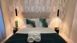 Una cama bien hecha con una colcha verde oscuro y ropa de cama blanca, dos almohadas decorativas con motivos naturales y dos lámparas de noche en mesitas a juego.