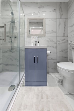 A bathroom with a glass-enclosed shower, a marble-tiled wall, a grey cabinet with two doors, and a toilet. - Harlesden Comfort Studio (Greater London)