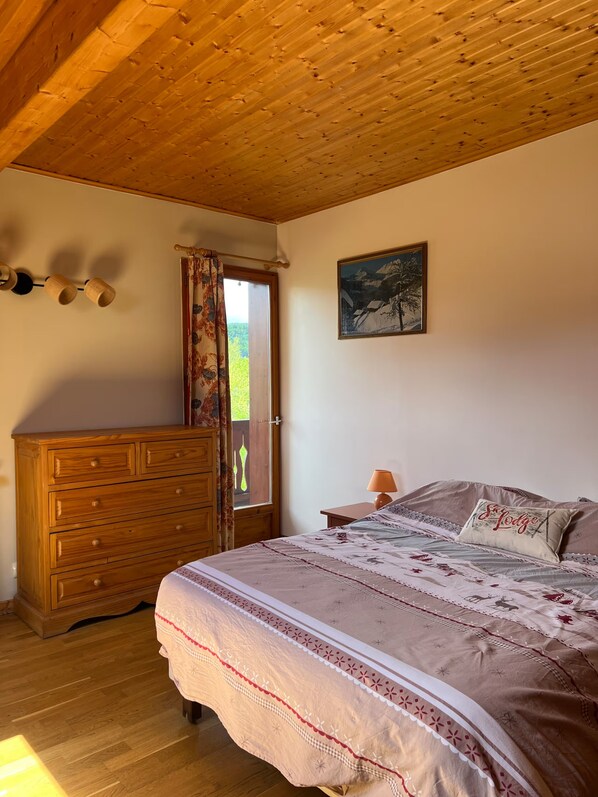 Une chambre avec un plafond en bois, un lit recouvert d’une couvre-lit à motifs, une commode en bois, une fenêtre avec des rideaux et un tableau accroché au mur.