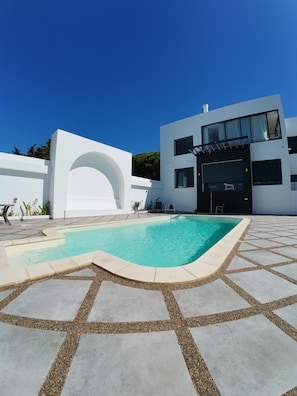 Un edificio moderno con piscina, una zona de estar exterior cubierta y un cielo azul despejado.