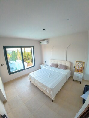 Ein Schlafzimmer mit einem großen Bett, einem Nachttisch, einer Kommode und einem Fenster mit Blick auf Bäume.