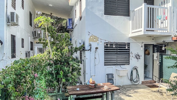 Un patio con mesa y bancos de madera, rodeado de follaje y un edificio blanco con varias ventanas y unidades de aire acondicionado.